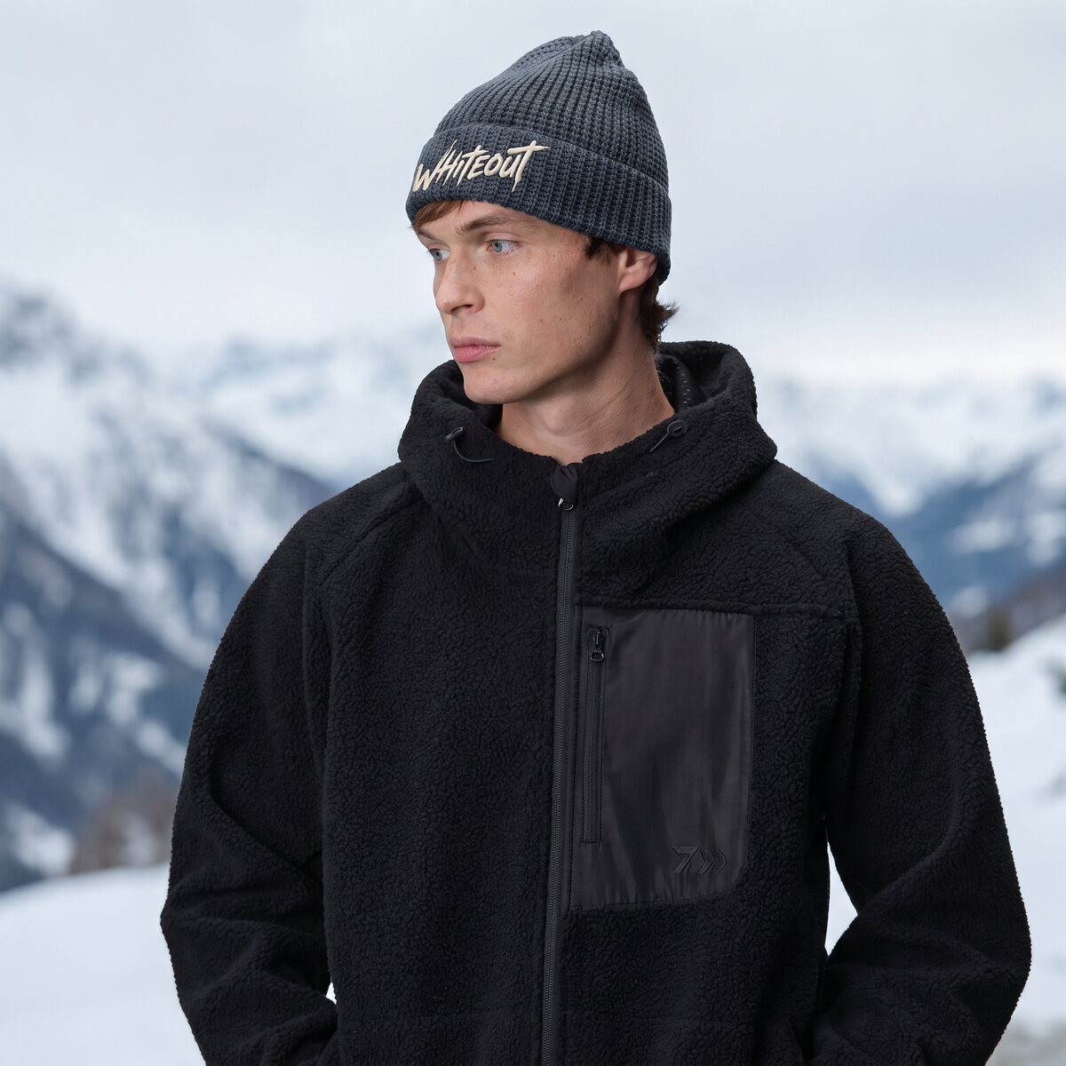 Model in a charcoal waffle beanie with the Whiteout script logo, snowy peaks behind